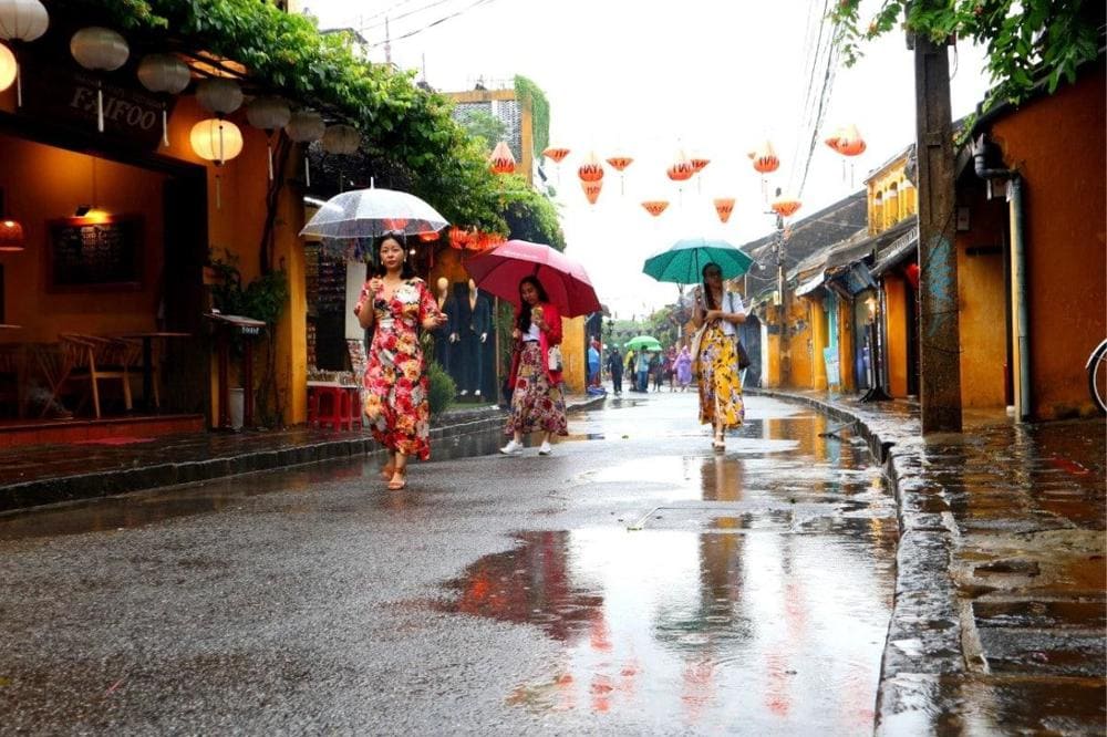 Locals and travelers take shelter under shop awnings as warm rain falls on a quiet Hoi An street